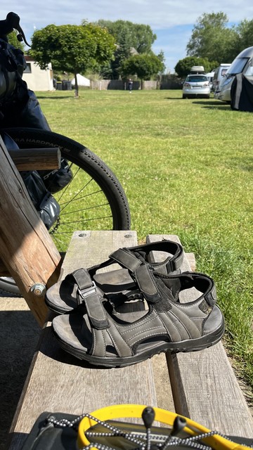 Ein Paar Sandalen auf einer Holzbank mit einem Fahrradrad auf der linken Seite und Campingfahrzeugen im Hintergrund auf einer Wiese.