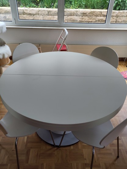 Ein runder weißer Tisch mit vier weißen Stühlen drum herum. In der Mitte des Tisches ist eine Fuge, die auf die Ausziehfunktion hinweist.