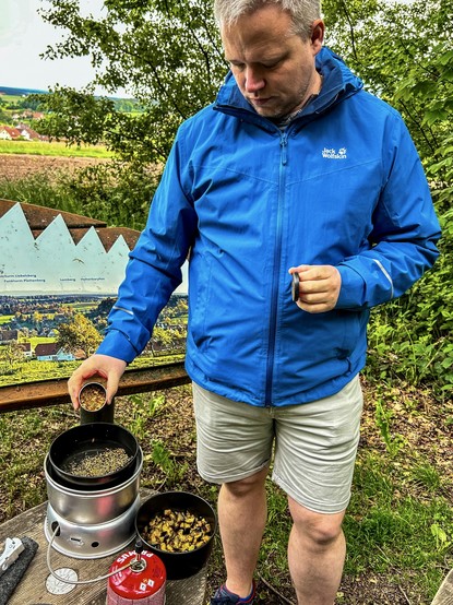 Mann mit blauer Regenjacke kocht mit einem Trangia-Kocher in der Natur