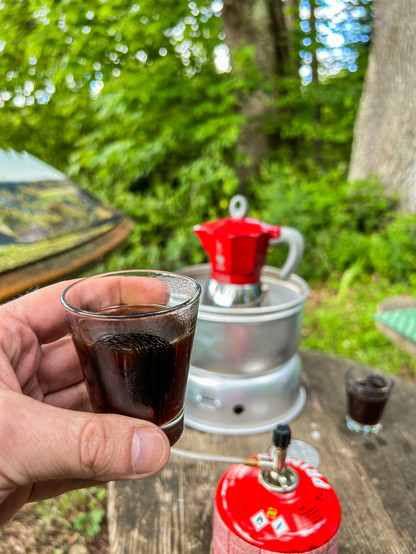 Espresso im Glas, dahinter rote Bialetti-Herdkanne in Trangia-Kocher