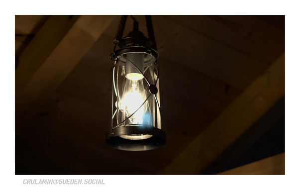 Einer alten Gaslaterne nachempfundene Leuchte mit LED-Glühbirne hängt in einem hölzernem Gebälk. Es ist dunkel, die Glühbirne ist an und verbreitet warmes Licht.