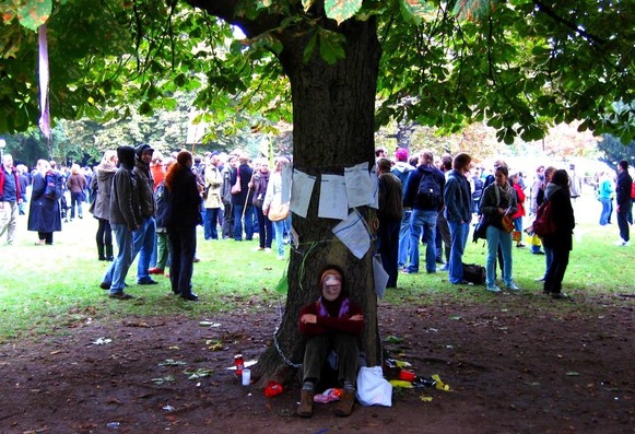 Frau sitzt angekettet an einem mit Flugblättern versehenen Baum - im Hintergrund Demonstranten im Park 