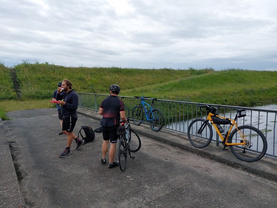 Mehrere Radfahrer stehen auf einer Brücke über einen Graben hinter dem Rheindamm. Ihre Räder lehnen teilweise am Geländer.