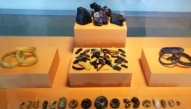 Museumsvitrine mit blau-schwarzen Glasbrocken, gelben und blauen Armreifen, Glasperlen und weitere Bruchstücke von Glasprodukten