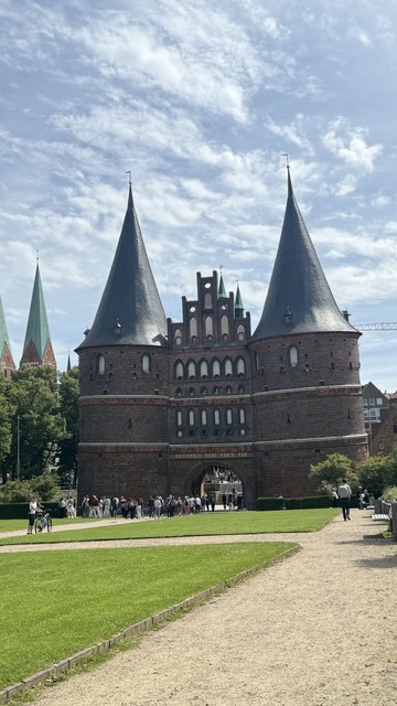 Das historische Holstentor mit zwei hohen, konischen Türmen und einem großen Torbogen am Eingang, umgeben von Grün und Menschen zu Fuß.