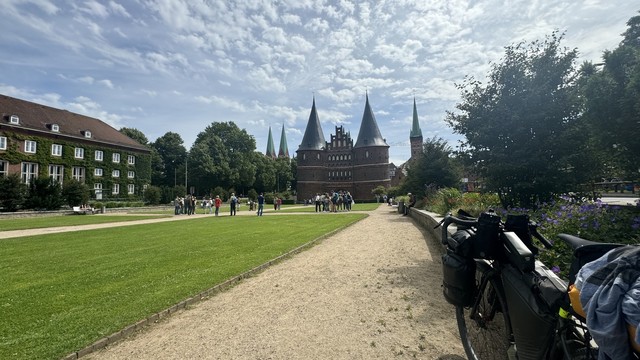 Das historische Holstentor in Lübeck mit mehreren Türmen, umgeben von Gruppen von Menschen auf einer Grasfläche. Ein Fahrrad ist auf einem Weg rechts geparkt, mit Blumensträuchern in der Nähe. Der Himmel oben ist teilweise bewölkt.