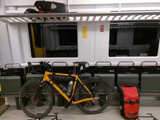 A yellow gravel bike with a removable pannier rack is tied to the wall of a train. One red pannier bag stands next to the bicycle on the floor, the other one and the helmet are in the luggage rack. Two bottles, a small saddle bag for tools and a spare tube and a small frame bag are mounted at the bicycle as well.