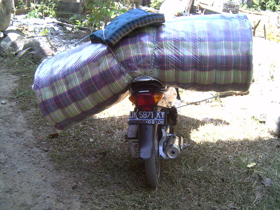 Matratzentransport in Indonesien. Eine zusammen gerollte Matratze liegt quer auf dem Sozius einen kleinen Motorrads.