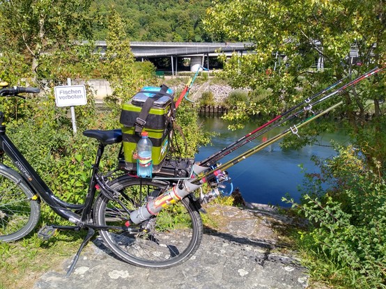 Ein Fluss, dahinter Gebüsch und eine Brücke. Im Vordergrund steht ein mit Anglermaterial vollgepacktes Fahrrad. Direkt dahinter ein Schild mit der Aufschrift 