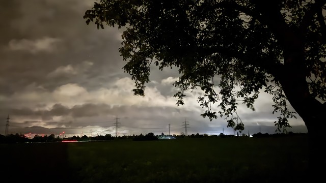 Eine nächtliche Landschaft mit einem dunklen Feld, einem Baum im Vordergrund und tief hängenden Wolken. Im Hintergrund sind schwache Lichter von entfernten Gebäuden und Stromleitungen gegen den bewölkten Himmel zu sehen.