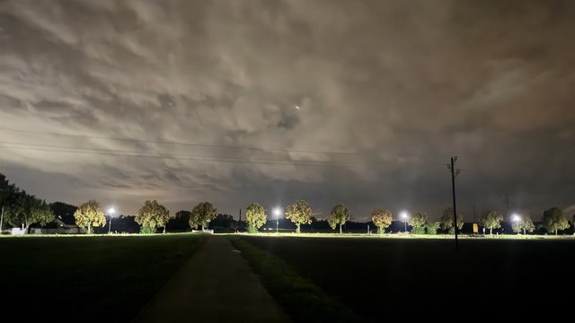 Eine Nachtszene mit einem Weg neben beleuchteten Bäumen und mehreren Lichtmasten unter einem bewölkten Himmel. Das Ambiente ist ruhig, die Bäume erscheinen in Silhouette gegen die dunklen Wolken.