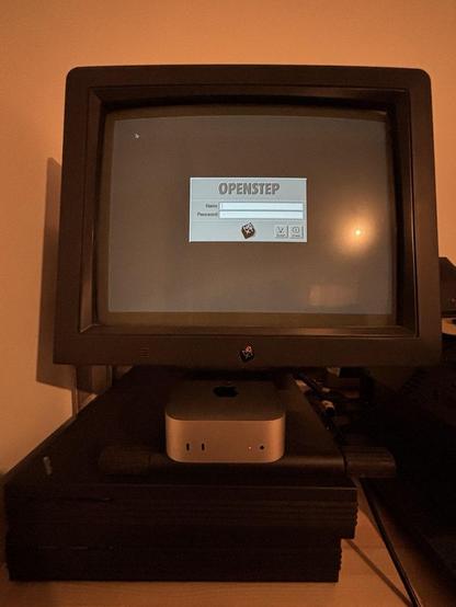 Two mono NeXTstations (one non-Turbo and one Turbo), one of them showing the OPENSTEP 4.2 login window on a grayscale Megapixel CRT. An M4 Mac Mini is sitting beneath the monitor.