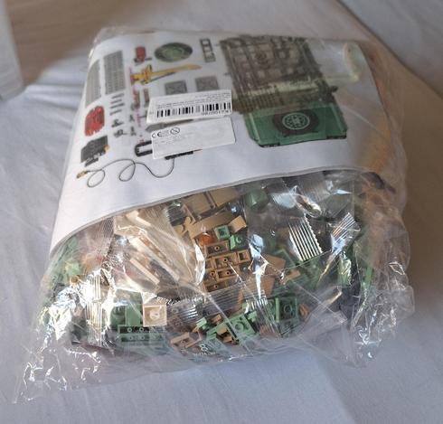 Das Bild zeigt ein ungeöffnetes Lego-Set, das in einer durchsichtigen Plastikverpackung auf einem weißen Untergrund liegt. Man sieht die vielen kleinen Lego-Steine in verschiedenen Beige- und Grüntönen. Darüber liegt die Anleitung mit Bildern des fertigen Modells, darunter ein grünes Fahrzeug mit einem Globus-Emblem, sowie Illustrationen der benötigten Teile. Ein weißer Aufkleber mit einem Barcode ist ebenfalls sichtbar.
