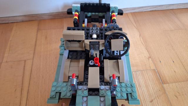 Das Bild zeigt einen Lego-Nachbau des Innenraums eines Autos, aufgenommen von oben. Das Lenkrad befindet sich rechts, und in der Mitte befindet sich ein Armaturenbrett mit verschiedenen Bedienelementen. Die Sitze sind beige und es gibt rote und graue Akzente. Der gesamte Aufbau besteht aus Legosteinen in verschiedenen Farben wie Schwarz, Beige, Grau und Grün. Der Hintergrund ist ein Holzboden.