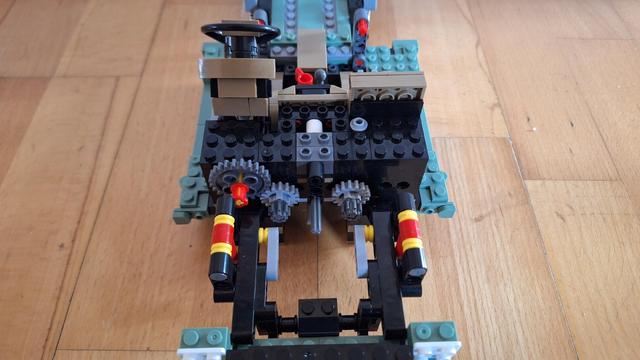 Das Bild zeigt das Fahrgestell eines Lego-Fahrzeugs auf einem Holzboden. Im vorderen Bereich befindet sich ein Cockpit mit Lenkrad, gefolgt von einem Motorblock, der mit Zahnrädern und Achsen versehen ist. Die Aufhängung ist durch Stoßdämpfer und Achsen dargestellt. Das gesamte Modell besteht aus verschiedenen Lego-Steinen in Farben wie Schwarz, Grau, Braun, Rot, Gelb und einigen grünen Akzenten.