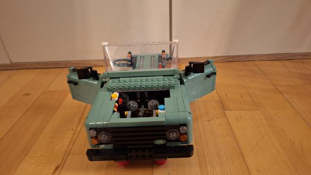 Das Bild zeigt ein Lego-Modell eines Land Rover Defender auf einem Holzboden. Die Farbe des Land Rovers ist ein helles Grün. Die Motorhaube des Wagens ist geöffnet und gibt den Blick auf ein kompliziertes Getriebe frei. Die Türen des Autos sind ebenfalls geöffnet. Man kann das Lenkrad und die Sitze im Innenraum sehen. Das Logo von Land Rover ist auf der Vorderseite des Fahrzeugs sichtbar. Der Hintergrund ist unscharf und zeigt eine helle Wand.