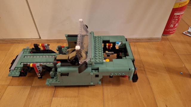 Das Bild zeigt ein unfertiges Lego-Modell eines grünen Geländewagens. Das Modell steht auf einem Holzboden vor einer weißen Wand. Der Wagen hat eine offene Fahrerkabine mit einem transparenten Windschutz. Die Karosserie ist größtenteils fertiggestellt, aber einige Teile, insbesondere an der Front, fehlen noch. Im Hintergrund steht eine Dose mit Aufschrift, die teilweise verdeckt ist.