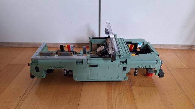 Das Bild zeigt ein Lego-Modell eines offenen Geländewagens, der auf einem Holzboden steht. Das Auto ist hellgrün und beige. Man sieht das detaillierte Interieur mit Sitzen, Lenkrad und Armaturenbrett. Am Heck des Wagens sind Werkzeuge befestigt. Im Hintergrund sind eine weiße Wand und ein Teil eines Stuhls zu sehen. Das Foto ist aus einer leicht erhöhten Perspektive aufgenommen.