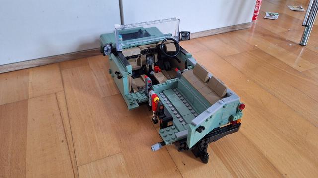 Das Bild zeigt das unvollständige Modell eines olivgrünen Land Rover Defender, das aus Legosteinen gebaut wurde. Das Modell ist ohne Karosserie, sodass das Interieur mit Lenkrad und Sitzen sichtbar ist. Die Aufhängung des Fahrzeugs ist ebenfalls deutlich erkennbar. Das Lego-Modell steht auf einem Holzboden vor einer weißen Wand.