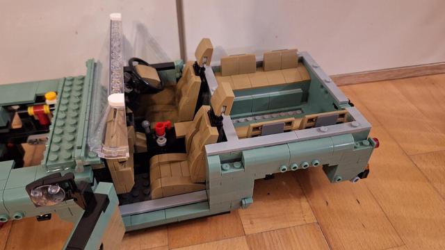 Das Bild zeigt einen unvollständigen hellgrünen Lego-Oldtimer. Zu sehen ist das Innere des Fahrzeugs mit hellbraunen Sitzen und einem schwarzen Lenkrad. Die Karosserie ist teilweise fertiggestellt, wobei einige Bereiche noch offen sind, was den Blick auf das darunter liegende Lego-Baumuster ermöglicht. Das Auto steht auf einem Holzboden.