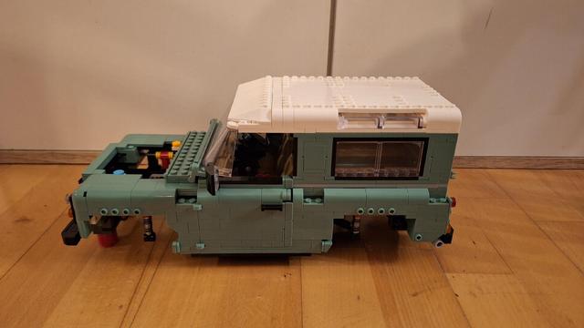 Das Bild zeigt ein unvollständiges Lego-Modell eines Land Rover Defender mit einem weißen Dach. Das Modell ist hauptsächlich in einem hellen Grün gehalten. Der vordere Teil des Fahrzeugs fehlt, sodass man in den Motorraum sehen kann. Der Rest des Fahrzeugs ist teilweise zusammengebaut, einschließlich der Karosserie, des Daches und einiger Innendetails. Das Modell steht auf einem Holzboden.