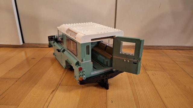 Das Bild zeigt ein Lego-Modell eines Land Rover Defender. Das Modell ist hellgrün und weiß und steht auf einem Holzboden. Die Heckklappe des Defenders ist geöffnet, sodass man den Innenraum mit Sitzen sehen kann. Das Modell ist sehr detailliert und sieht realistisch aus.
