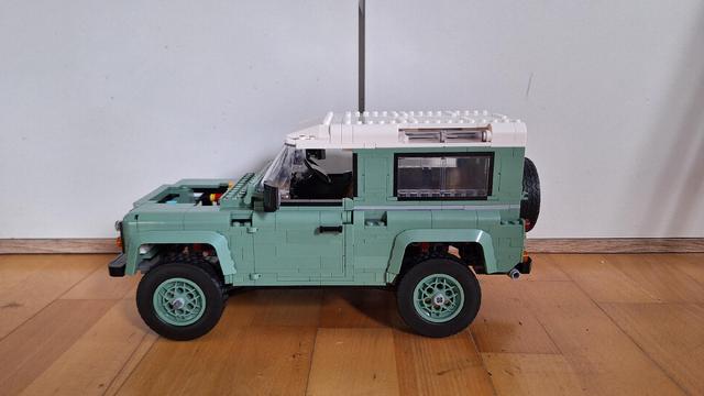 Das Bild zeigt ein Lego-Modell eines Land Rover Defender, aufgenommen aus einer leicht erhöhten Perspektive von hinten. Das Modell ist in einem hellen Grün gehalten, mit einem weißen Dach. Auf der Rückseite des Fahrzeugs befindet sich ein montiertes Reserverad und ein roter Feuerlöscher. Das Modell steht auf einem Holzboden.
