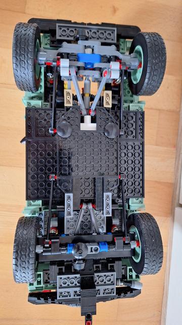 Das Bild zeigt die Unterseite eines Lego-Modellautos, das auf einem hölzernen Hintergrund steht. Die Karosserie des Autos ist größtenteils schwarz und olivgrün. Die Räder sind groß und haben ein grobes Profil. Man sieht die komplizierte Aufhängung und den Rahmen des Autos, bestehend aus grauen, blauen und roten Lego-Technic-Teilen. Die Konstruktion wirkt detailliert und technisch anspruchsvoll.