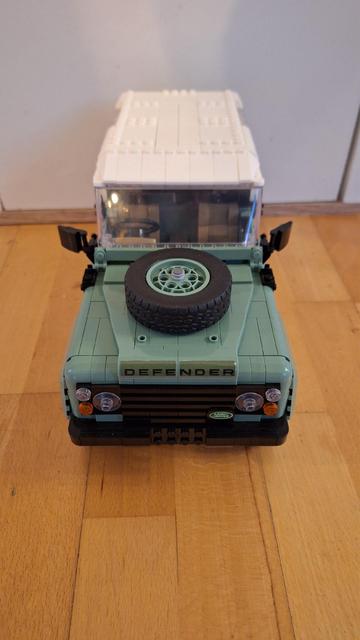 Ein Foto von vorne zeigt ein Lego-Modell eines hellgrünen Land Rover Defender. Auf der Motorhaube des Autos befindet sich ein Reservereifen. Der Wagen hat ein weißes Dach. Der Hintergrund ist unscharf, aber man kann eine weiße Wand und einen Holzboden erkennen.