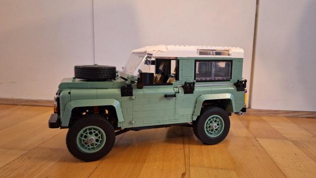 Ein Lego-Modell eines grünen Land Rover Defender steht auf einem Holzboden. Das Dach des Defenders ist weiß. Auf der Motorhaube ist ein Reserverad befestigt. Das Modell ist detailliert und zeigt die typische kastenförmige Form des Defenders. Der Hintergrund ist eine schlichte weiße Wand