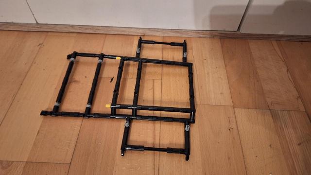 Auf einem Holzboden liegt ein Konstrukt aus schwarzen Kunststoffrohren und Verbindungsstücken. Es besteht aus drei rechteckigen Rahmen, die miteinander verbunden sind. Zwei der Rahmen sind übereinander gestapelt, während der dritte Rahmen seitlich daran befestigt ist. Einige gelbe und graue Verbindungsstücke sind sichtbar. Im Hintergrund befindet sich eine weiße Wand.

Bereitgestellt von @altbot, generiert mit Gemini