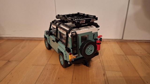 Ein Foto aus der Froschperspektive zeigt einen hellgrünen Lego Land Rover Defender auf einem Holzboden. Der Defender hat ein schwarzes Gepäckträger auf dem weißen Dach. Am Heck befindet sich ein Reserverad und ein roter Feuerlöscher. Das Nummernschild am Heck des Fahrzeugs lautet "L3 HUE". Im Hintergrund befindet sich eine weiße Wand.
