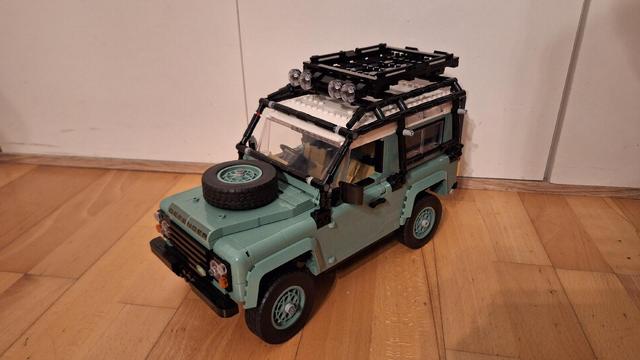 Das Bild zeigt ein Lego-Modell eines Land Rover Defender in einem hellen Grünton. Das Modell steht auf einem Holzboden vor einer weißen Wand. Auf der Motorhaube befindet sich ein Reserverad. Das Dach des Defenders ist weiß, und darauf ist ein schwarzer Gepäckträger mit Scheinwerfern montiert. Das Modell ist detailliert und wirkt realistisch.