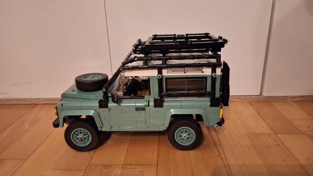 Ein Lego-Modell eines hellgrünen Land Rover Defender steht auf einem Holzboden vor einer weißen Wand. Das Modell hat ein schwarzes Dachgestell und ein Reserverad auf der Motorhaube. Die Räder sind ebenfalls hellgrün mit schwarzen Reifen. Das Modell ist detailliert und realistisch gestaltet.