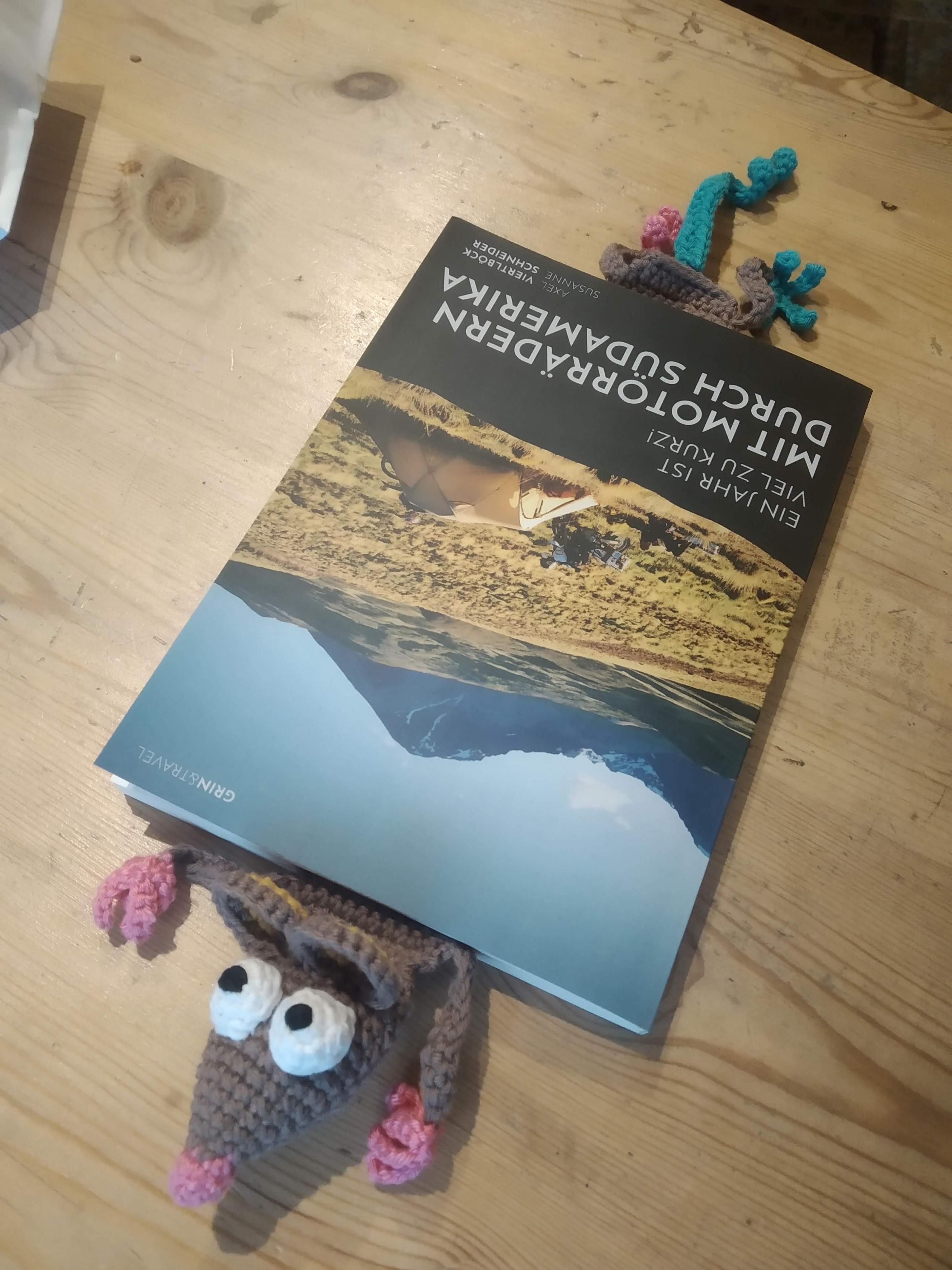Eine gehäkelte Ratte als Lesezeichen liegt in einem Buch. Vorne schaut der Kopf und die Vorderpfoten raus, oben die Hinterbeine und der Schwanz