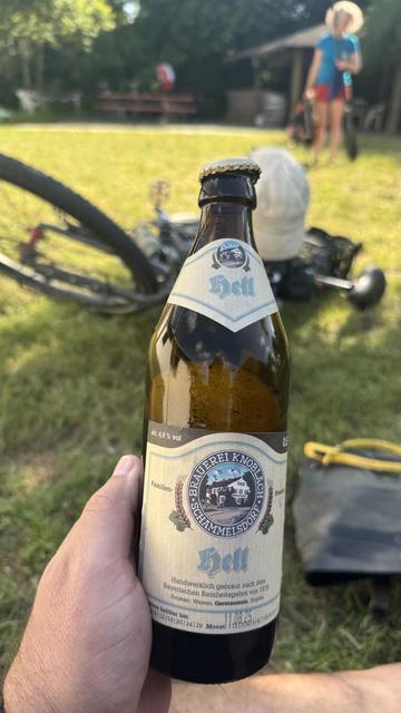 Eine Hand hält eine Flasche Bier mit der Aufschrift "Hell", mit einem Etikett, das darauf hinweist, dass sie nach dem bayerischen Reinheitsgesetz hergestellt wurde. Im Hintergrund steht ein Fahrrad und eine Person in der Nähe eines schattigen Bereichs, umgeben von Gras.