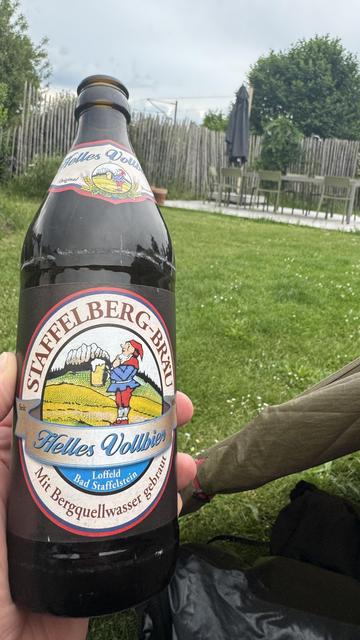 Eine Hand hält eine Flasche Staffelberg-Bräu Helles Vollbier mit einem bunten Etikett. Der Hintergrund zeigt eine grasbewachsene Fläche mit Gartenmöbeln und einem Zaun, unter einem bewölkten Himmel.
