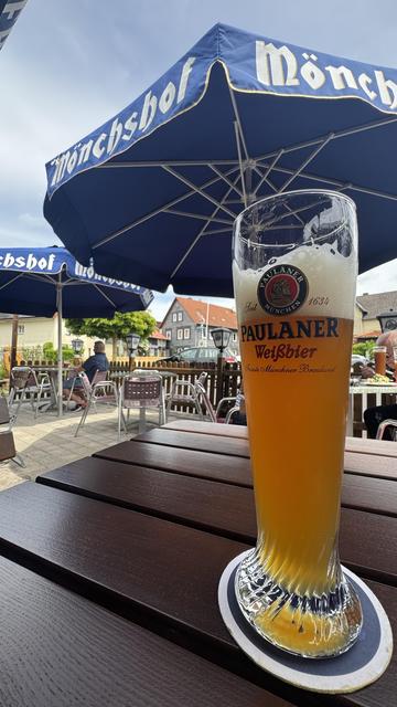 Ein Glas Paulaner Weißbier ist prominent auf einem Holztisch ausgestellt, mit einem Regenschirm mit dem Mönchshof-Logo im Hintergrund. Die Szene zeigt einen Sitzbereich im Freien mit Kunden in der Ferne und einem klaren Himmel.