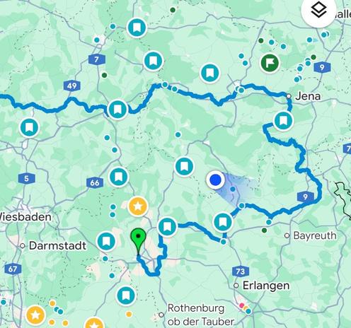 Eine Karte, die eine gewundene blaue Route durch eine grüne Landschaft mit verschiedenen Symbolen zeigt, die Sehenswürdigkeiten anzeigen, einschließlich Markierungen für Campingplätze, Attraktionen und andere Orte in der Nähe von Jena, Erlangen und Darmstadt.