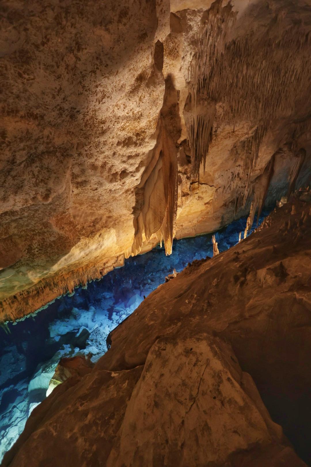 Tropfsteinhöhle mit Wasser. Aufgenommen im Inneren der Höhle von einem erhöhten Standort. Von oben geht der Blick auf klares Wasser, welches durch Kunstlicht beleuchtet ist. Die Felsen sind in warmem Licht, während das Wasser blau leuchtet.