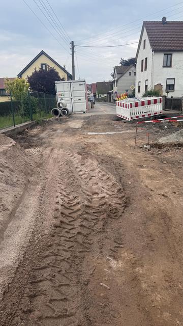 Eine Baustelle mit einer unbefestigten Straße mit Reifenspuren, einem großen Container und Verkehrsschranken. Im Hintergrund gibt es Wohngebäude und Grün. Das Gebiet scheint sich in der Entwicklung oder Reparatur zu befinden.