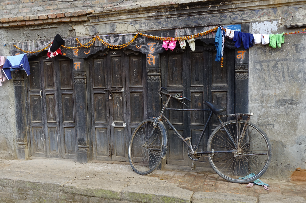 Alte Hauswand aus bröselnden Backsteinen, alles etwas Grau und schmutzig. Drei dunkle hölzerne verschlossene Türöffnungen mit geschnitzten Türstöcken, orangene Blumengirlande darüber an der einige bunte Wäschestücke zum trocknen hängen.
Davor ein altes Schwarzes Fahrrad an die Wand gelehnt