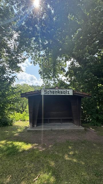 Ein hölzerner Unterstand mit dem Namen "Schenksolz" befindet sich in einer grünen Fläche, umgeben von Bäumen. Sonnenlicht filtert durch das Laub und schafft eine ruhige Atmosphäre.