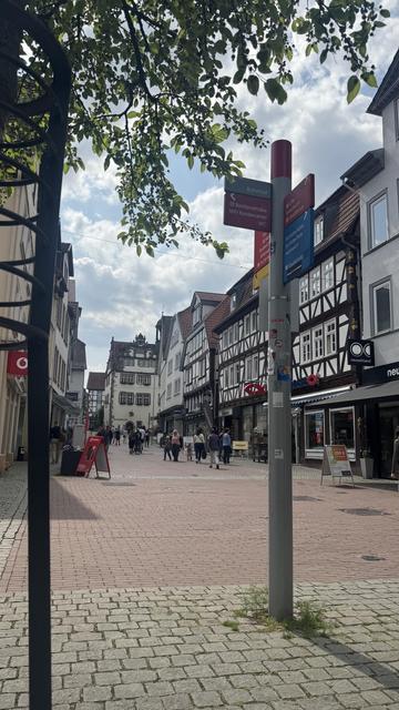 Eine malerische Straßenszene mit einem gepflasterten Gehweg, der von Fachwerkhäusern gesäumt ist. Fußgänger schlendern vorbei, und Wegweiser weisen auf nahe gelegene Sehenswürdigkeiten hin. Der Himmel ist teilweise bewölkt, mit grünen Baumblättern im Vordergrund.