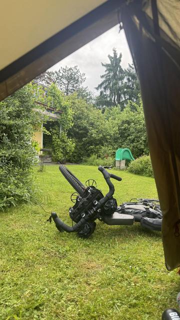 Blick aus dem Inneren eines Zeltes, das ein Fahrrad zeigt, das auf dem Gras liegt. Es gibt üppig grüne Pflanzen und Bäume im Hintergrund, zusammen mit einem gelben Gebäude und einer grünen Rutsche. Der Himmel ist bewölkt.