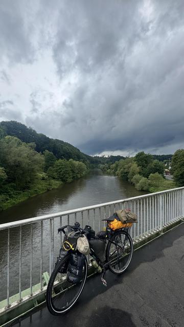 Ein Fahrrad ist auf einer Brücke mit Blick auf einen Fluss geparkt, der von üppigem Grün umgeben ist. Der Himmel ist mit dunklen Wolken bedeckt, was auf Regenpotenzial hindeutet.