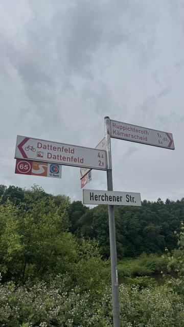 Ein Wegweiser, der Entfernungen zu verschiedenen Orten wie Datenfeld, Ruppichteroth und Kämerscheid anzeigt. Die Landschaft zeigt Grün und einen bewölkten Himmel.
