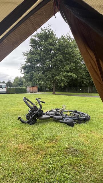 Eine Ansicht aus dem Inneren eines Zeltes zeigt eine grasbewachsene Fläche mit einem umgestürzten Fahrrad im Vordergrund, einem Baum im Hintergrund und einem links geparkten Wohnmobil. Der Himmel ist bedeckt.
