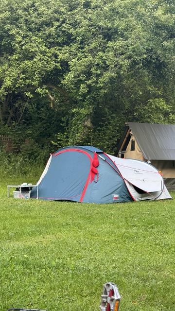 Ein blau-graues Zelt auf grünem Gras, umgeben von Bäumen. Ein kleiner Tisch mit Gegenständen steht in der Nähe, und ein weiteres Zelt ist im Hintergrund zu sehen.

An dem Zelt hängt ein roter BH. 