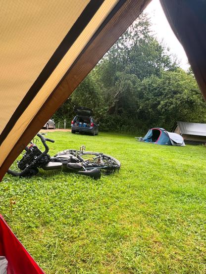 Ein Blick aus dem Inneren eines Zeltes, das grasbewachsene Campingplätze, ein blaues Auto mit offenem Kofferraum, Campingausrüstung auf dem Gras und zwei Zelte im Hintergrund unter einem baumdichten Gebiet zeigt.